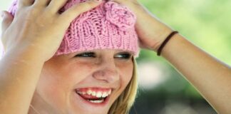 Niña rubia con sombrero de lana rosa, las manos en la cabeza y sonriendo.
