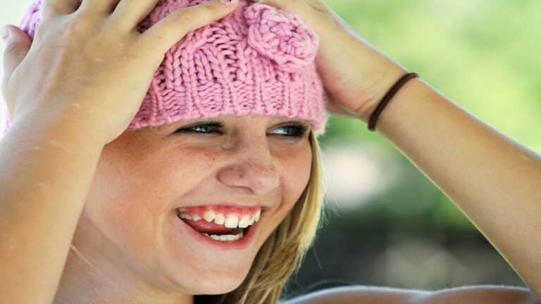 Niña rubia con sombrero de lana rosa, las manos en la cabeza y sonriendo.